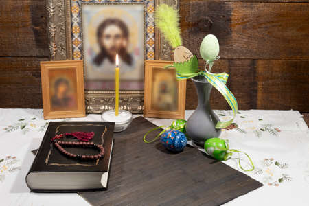 On the table in the background are icons. There is a burning candle, a Bible book with a rosary,...