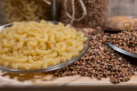 Buckwheat is scattered on a wooden board. Nearby is a transparent plate with pasta.
