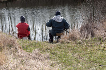 On the lake, a fisherman with a fishing rod catches fish. The boy sits next to him and looks.の写真素材