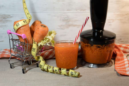 In the shopping trolley is a carrot and a centimeter. Nearby is a glass of juice. Carrot puree is prepared in a blender.の写真素材