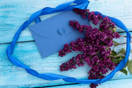On a blue plank background lies a blue envelope and a branch of purple lilac. The composition is framed by a ribbon.の写真素材