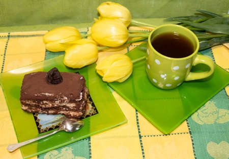 On the table is a cup of tea and a plate with a slice of cake and a spoon. Nearby are yellow tulips.の写真素材