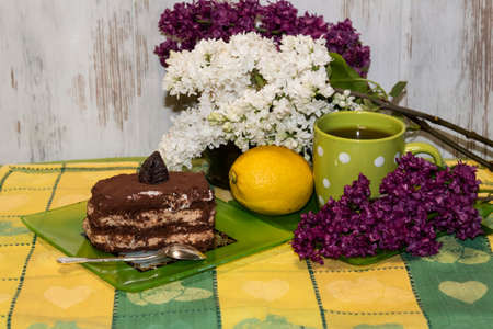 On the table is a cup of tea and a plate with a slice of cake and a spoon. Lying lemon and branches of lilac white and purple.の写真素材