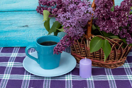 On a blue background is a basket with purple lilacs. Nearby is a cup of tea and a candle.の写真素材