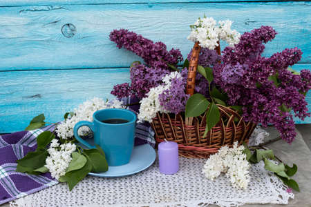 On a white knitted napkin is a cup of tea. Nearby is a bouquet of white and purple lilacs in a basket and a candle.の写真素材