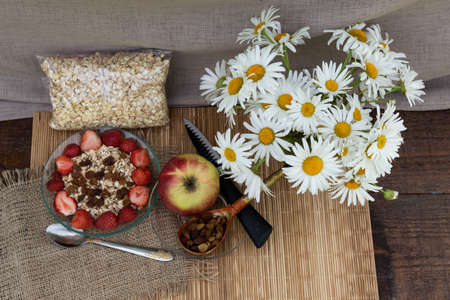 On the table is a bouquet of daisies, an apple is lying, raisins in a wooden spoon. A bowl of oatmeal muesli with strawberries is cooked in a bowl.の写真素材