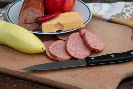 On the board lies sliced pieces of sausage and yellow pepper. On the plate you can see hot pepper,...