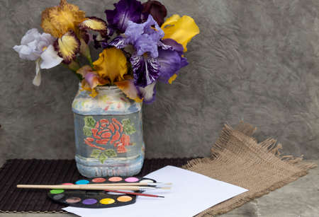 On the table is a bouquet of colorful irises. Nearby lies a sheet of paper and a palette with paints.の写真素材