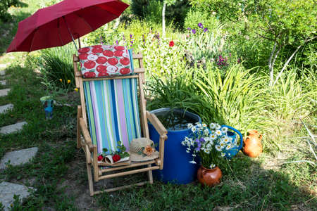 Next to the water barrel is a deck chair under a red umbrella. In a chaise lounge there is a female hat, an open book with roses. Nearby stands in a clay pot a bouquet of daisies.の写真素材