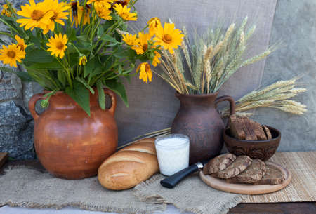 On the table is a bouquet of yellow flowers in a clay pot. Nearby lies white bread, stands a glass of milk, lies a knife and sliced brown bread. In the background is a jug with ears of wheat.の写真素材