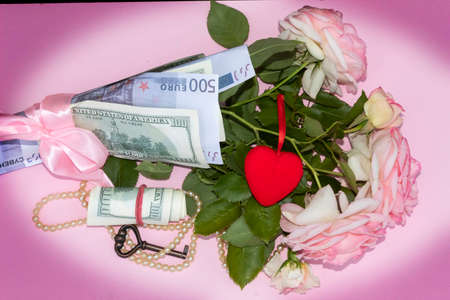 View from above. On a pink background with darkened corners lies a bouquet of roses wrapped in banknotes and a red heart. Nearby are beads, a key and a bundle of money.の写真素材