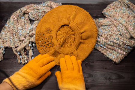 Two female hands in yellow woolen gloves lie on a knitted beret. Nearby lies a beige scarf.