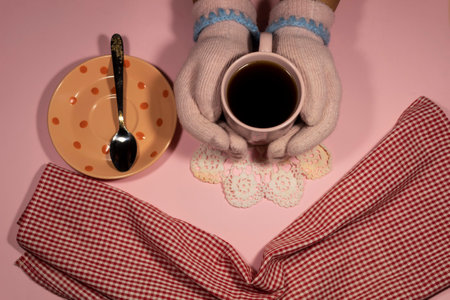 Two hands in pink knitted gloves hold a cup of coffee. Nearby is a saucer with a spoon and a napkin.