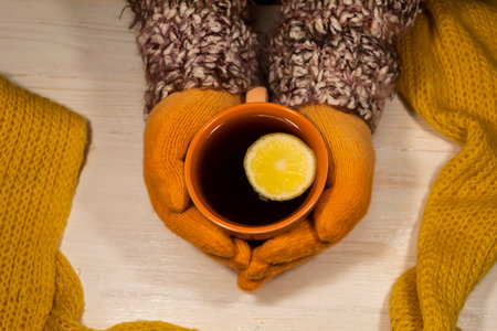 On a white table, gloved hands hold an orange cup of coffee with lemon. Nearby is a knitted woolen...