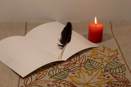 A red burning candle stands on an embroidered linen tablecloth, a sheet of white paper and a bird's...