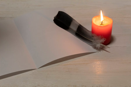 There is a red burning candle on a white table, a sheet of white paper and a feather of a bird.の写真素材