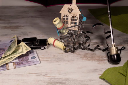 Under the umbrella is a supermarket trolley with a mock-up of a house and money. Nearby there is a toy car with money and three metal figures.の写真素材