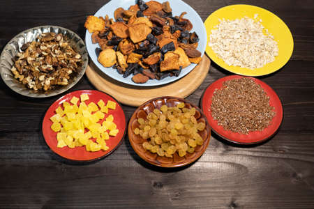 Dried fruits, oatmeal, flax seed, raisins, candied pineapple and walnuts lie on a dark background on multi-colored saucers.の写真素材
