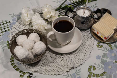 View from above. There are white carnations on a white tablecloth, a cup of coffee on a white saucer, a sugar bowl with sugar, a candy bowl and a saucer of cakes.の写真素材