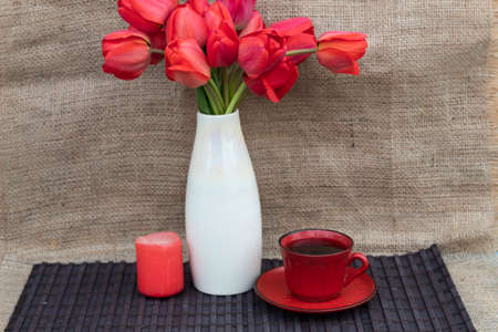 There is a white vase with red tulips, a candle and a red cup of coffee on a dark wooden napkin.の写真素材