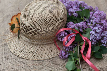 A woman's hat with a flower lies on a jute fabric. Nearby are lilac flowers with a red ribbon.の写真素材