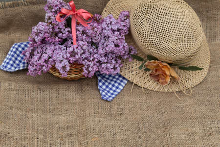 A woman's hat with a flower lies on a jute fabric. Nearby is a basket with lilac flowers and a red bow.の写真素材