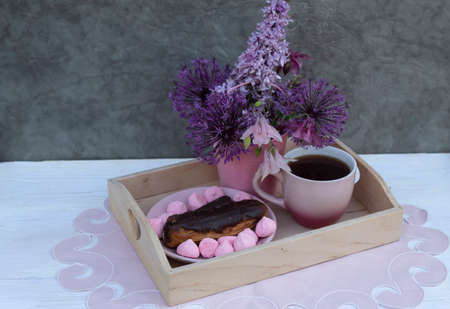 There is a cup of coffee on a tray, a dessert on a saucer. There is a pink bucket of flowers nearby.