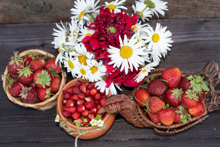 View from above. On a wooden background there is a pot with wild strawberries, wicker vases with strawberries and a bouquet of daisies and poppies.の写真素材