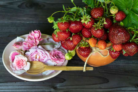 View from above. On a dark background, there is a bouquet of red ripe strawberries and a plate with a pink rose and a wooden spoon.の写真素材