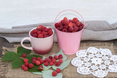 There is a cup and a bucket of raspberries on a jute fabric. Berries also lie on the raspberry leaves nearby.の写真素材