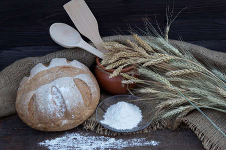 On the table is an earthen pot with a wooden spoon and spatula, a plate of flour, ears of wheat and a loaf of bread.の写真素材