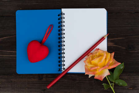 On a dark wooden background lies an open notebook, on one page of which is a red heart, and on the other - a rose and a red pencil.の写真素材