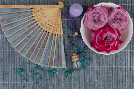 On a light wooden background there is sea salt, aromatic oil, a candle, open fan and a plate with floating roses.の写真素材