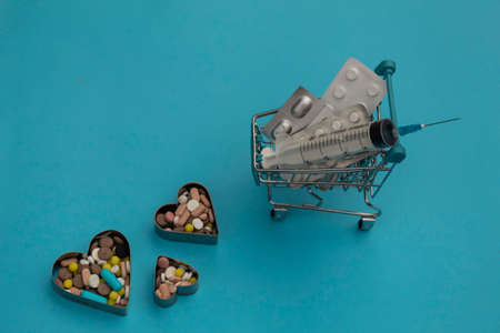 A cart with plates of pills and a syringe stands on a blue background. Nearby in the heart farms are multi-colored pills.の写真素材