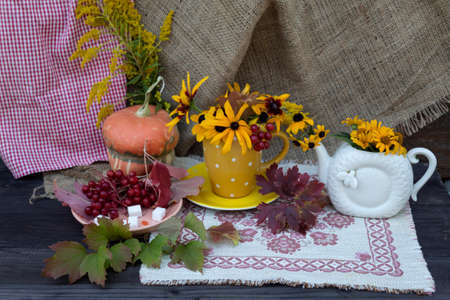 There is a teapot with yellow flowers on a napkin, a cup on a saucer with flowers, a saucer with viburnum and sugar cubes. Behind there is a pumpkin that looks like a mushroom.の写真素材