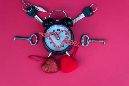 An old alarm clock with bells lies on a red background. There are keys and two red hearts next to it.の写真素材