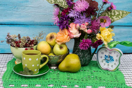 Still life. On the table is a bright bouquet of autumn flowers, a clock, a vase of apples, and a pear. Nearby is a cup of mint tea. Behind is a bowl with viburnum berries, mint and other herbs.の写真素材