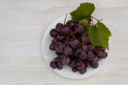 On a white wooden background, a ripe bunch of purple grapes and grape leaves lie on a plate.の写真素材