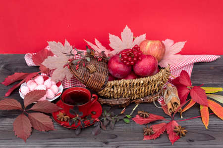 The original wicker vase contains red apples and viburnum berries. Nearby there is a cup of tea, a saucer of sweets, a branch of barberry, cinnamon sticks and anise stars, autumn leaves are scattered.の写真素材