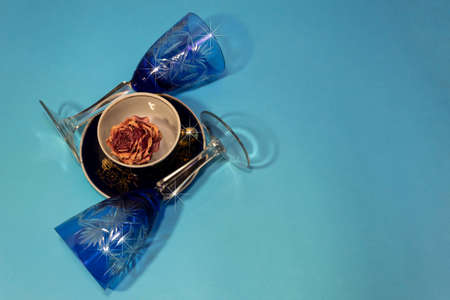 Against a blue background, there are two blue glasses and a cup on a saucer with a dry rose flower.