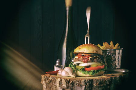 Fork in burger with mushrooms, tomatoes, cucumbers, salat, cheese and onions on a wooden base with fries, oil and garlic, retroの写真素材