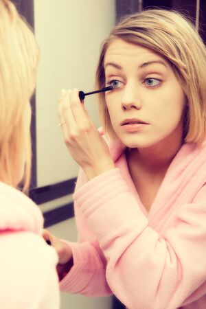 Blonde woman wearing in dressing-gown applying mascara in mirror at home.の写真素材