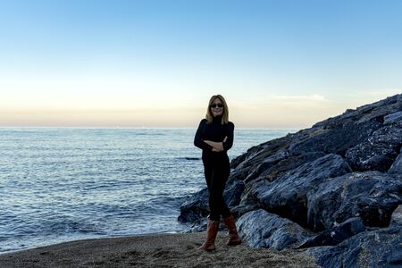 blonde woman with over 50, dressed in pants and black shirt and with cap, posing with the sea in the backgroundの写真素材