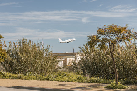 White passenger airplane taking off over a small building and some bushes and trees, on a sunny day...