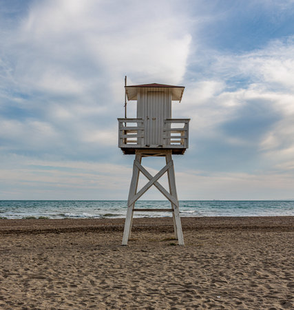Empty white lifeguard tower stands on a sandy beach, overlooking a calm sea under a cloudy skyの写真素材