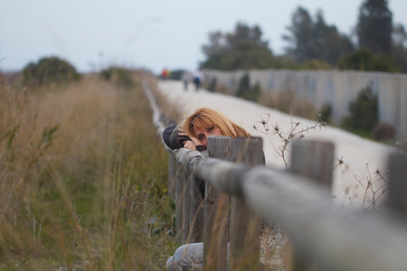 Woman with blonde hair resting on a wooden fence, looking towards the camera with a pensive expressionの写真素材