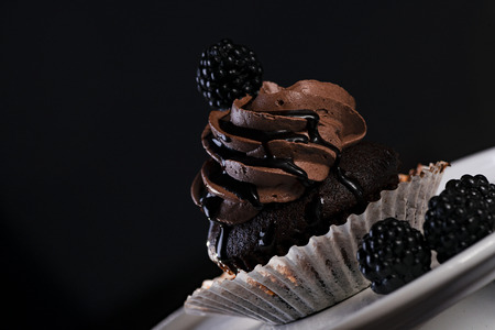 One chocolate cupcake with chocolate cream and liquid chocolate  on white cake stand and dark background. Decorated with blackberryの写真素材