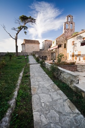 An empty narrow alley in the Old Town of Budva, Montenegro. Green grass, flowers, old tree and blue sky above.の写真素材