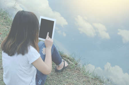 Young woman sitting near lake, sky reflection and using tablet , Vintage color styleの写真素材