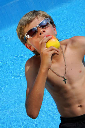 a 10-years old American - German boy with sunglasses sitting at a swimming pool and enjoying eating a delicious apple with closed eyes in the summer sunの写真素材